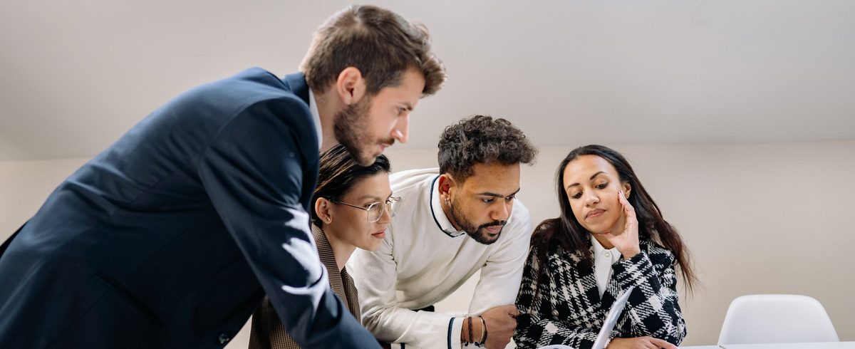 Four colleagues in a bright office lean over a laptop and review a proposal together.